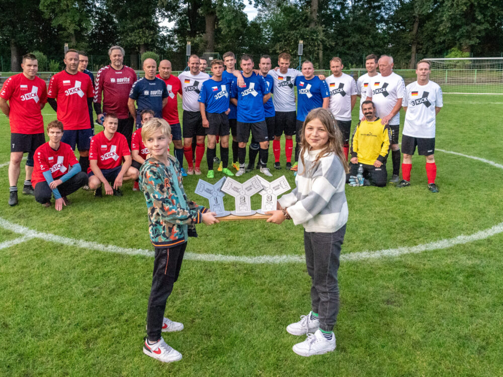 Zwei Kinder halten drei Pokale in der Hand. Im Hintergrund Fußballspieler auf dem Rasen als Gruppenfoto in unterschiedlichen Trikots.