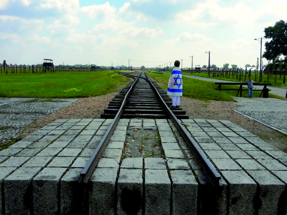 Ein Kind steht eingehüllt in eine Israel-Flagge an einem Bahngleis in der Gedenkstätte Auschwitz-Birkenau.