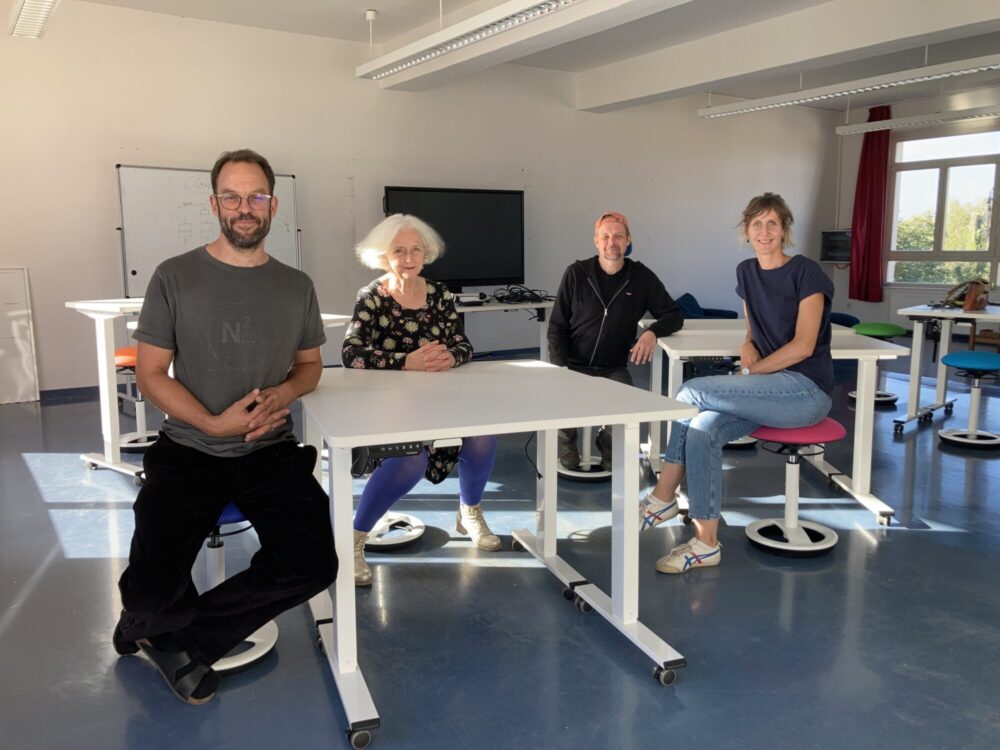 Vier Leute sitzen in einem Unterrichtsraum an weißen Tischen auf ergonomischen Hockern.