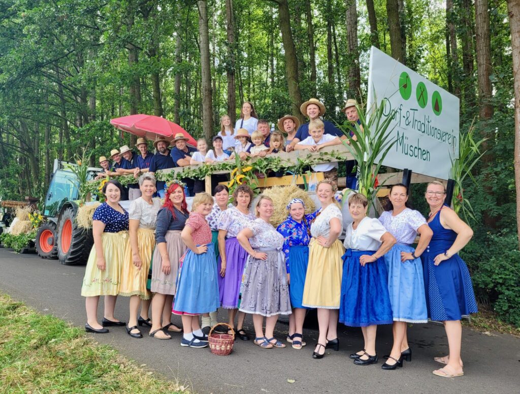 Frauen in Trachtenkleidung stehen vor einem Anhänger mit Traktor, Männer und Kinder mit Hüten stehen in der Gruppe auf dem Anhänger.