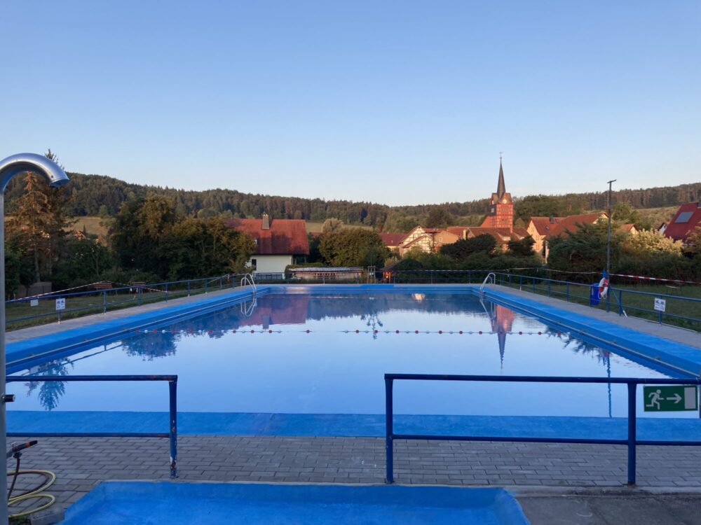 Blaues Becken eines Freibads, im Hintergrund Dorf und Kirche Rippershausen.