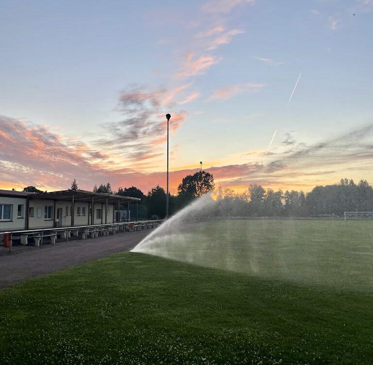 Bewässerung Sportplatz Prösen. Vereinshaus darüber Wolken im Sonnenuntergang.