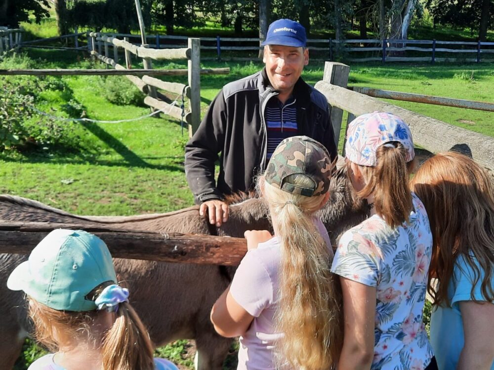 Kinder streicheln durch einen Zaun hindurch einen Esel. Der Tierpfleger steht neben dem Tier und lächelt in die Kamera.