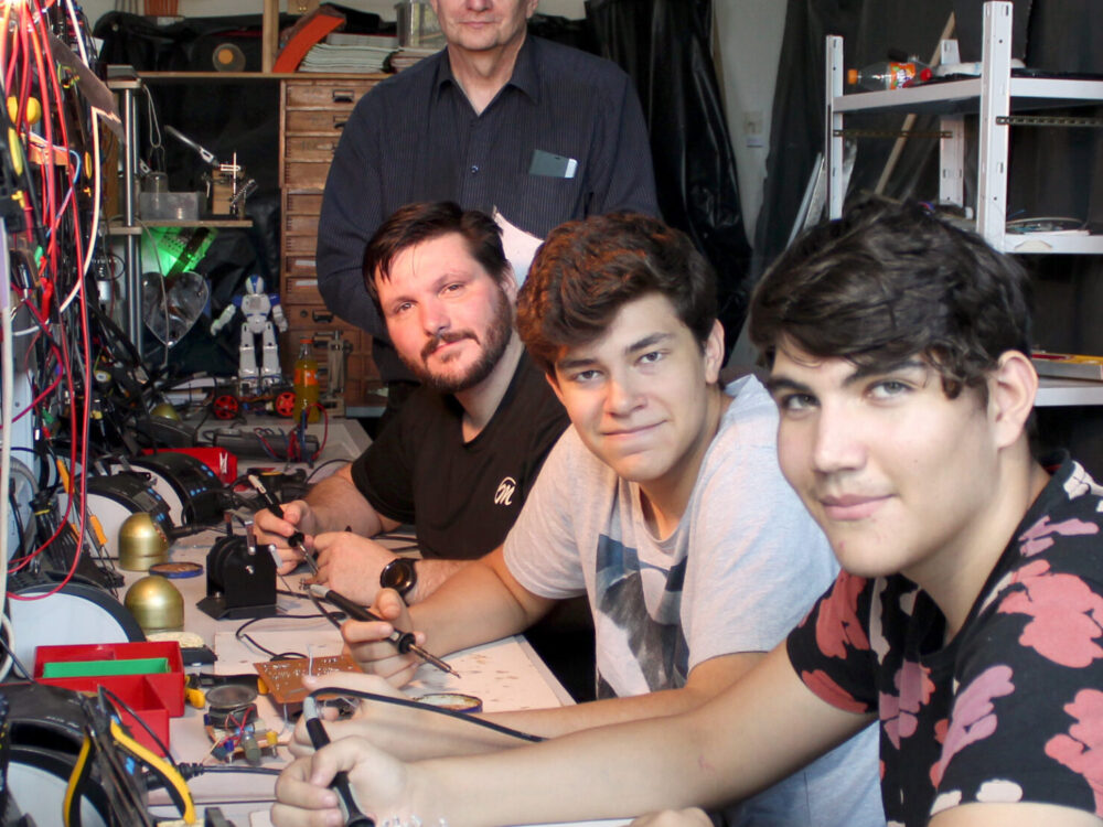 Jugendliche und Männer in einer Elektronikwerkstatt mit Bauteilen vor sich auf dem Tisch.