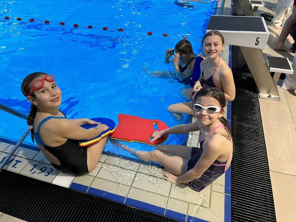 Mädchen in Schwimmausrüstung und Schwimmbrettern sitzen am Beckenrand eines Schwimmbeckens.