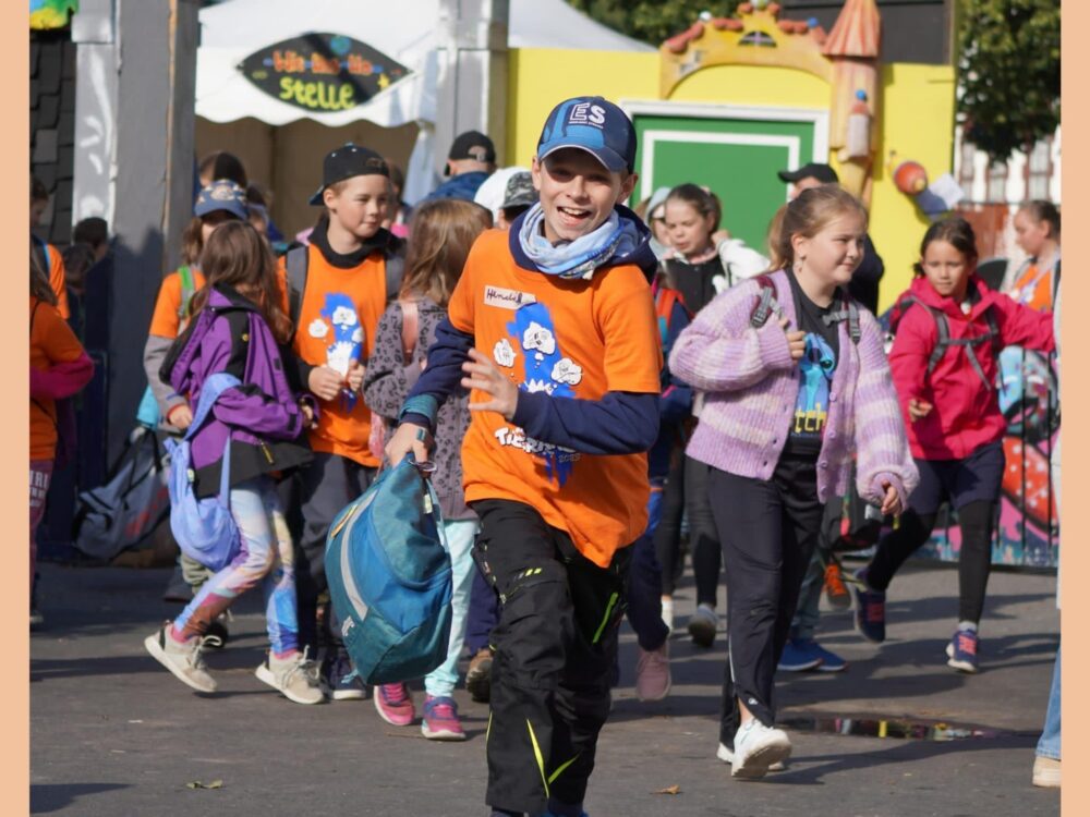 Ein Junge mit orangem T-Shirt rennt in Richtung Kamera, im Hintergrund weitere Kinder in bunter Kleidung.
