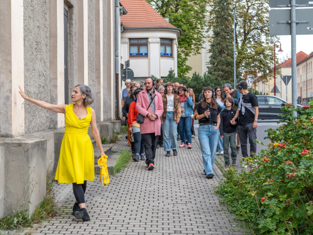 Eine Frau im gelben Kleid zeigt mit der Hand auf eine Fassade, Menschengruppe mit Kopfhörern folgt ihr auf dem Fußweg.