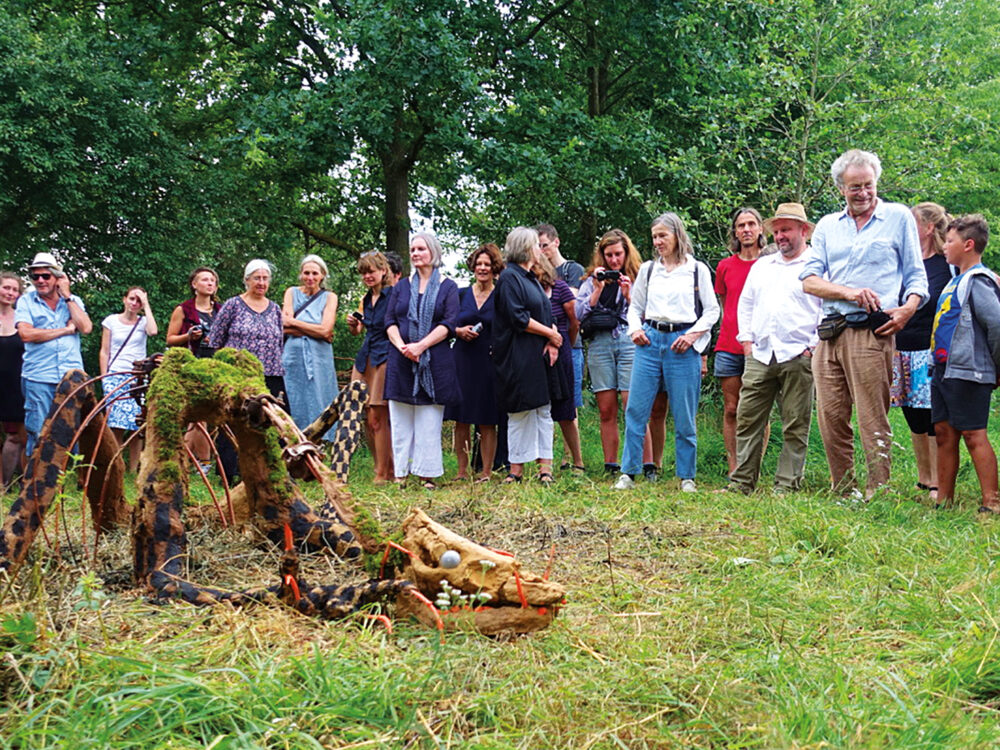 Menschengruppe betrachtet auf einer Wiese eine Skulptur aus Naturmaterialen.