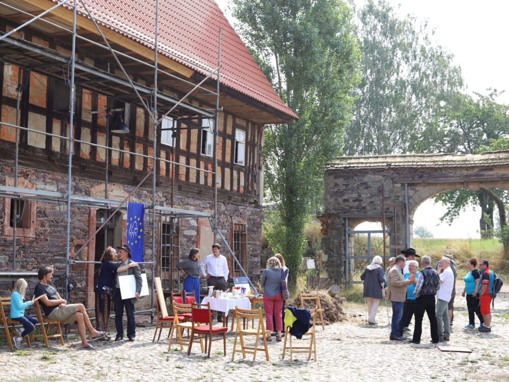 An einem sommerlichen Tag stehen Leute vor einem eingerüsteten Fachwerkhaus und Hoftor, Stühle und ein Tisch sind aufgebaut.