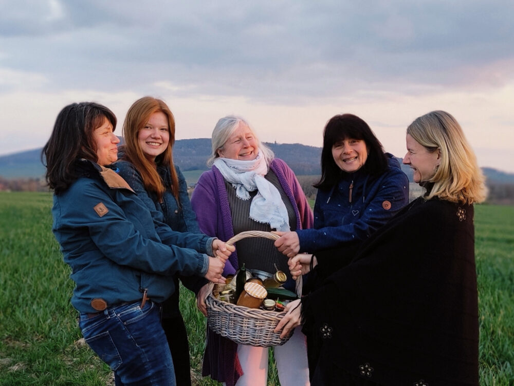 Fünf Frauen stehen auf einem Feld und halten einen Korb mit Einweggläsern und Gemüse in ihrer Mitte, im Hintergrund ein Berg.