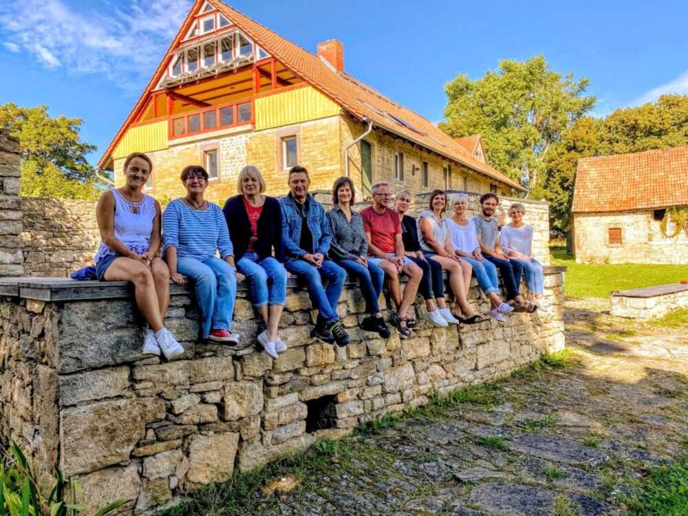 Menschen sitzen fröhlich auf einer Mauer vor einem restaurierten Bauernhaus.