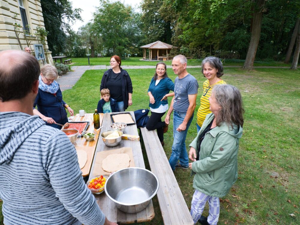 Mehrere fröhliche Personen stehen vor einem Haus um einen Tisch herum und belegen Pizzen auf Backblechen.