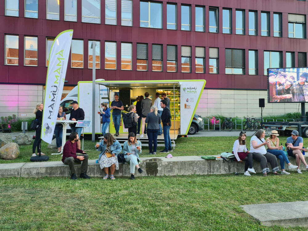 Bus mit Infostand vor einem Haus mit magentafarbener Fassade, davor sitzen und stehen Leute.