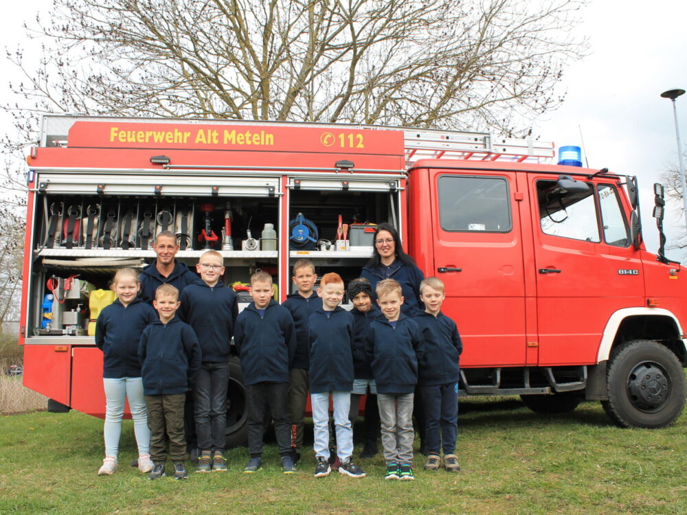 Kinder und zwei Erwachsene stehen als Gruppe vor einem an der Seite geöffnetem Feuerwehrauto.