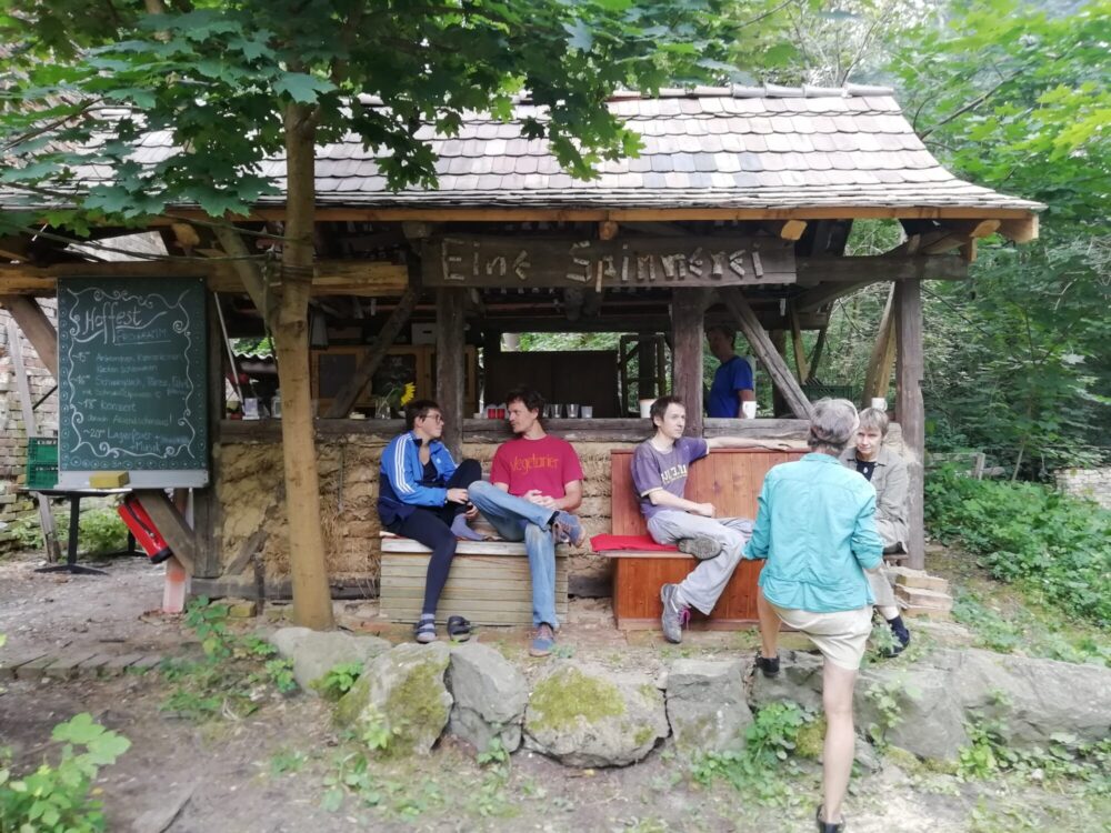 Vier leger gekleidete Leute sitzen vor einem rustikalen Ausschank im Grünen.