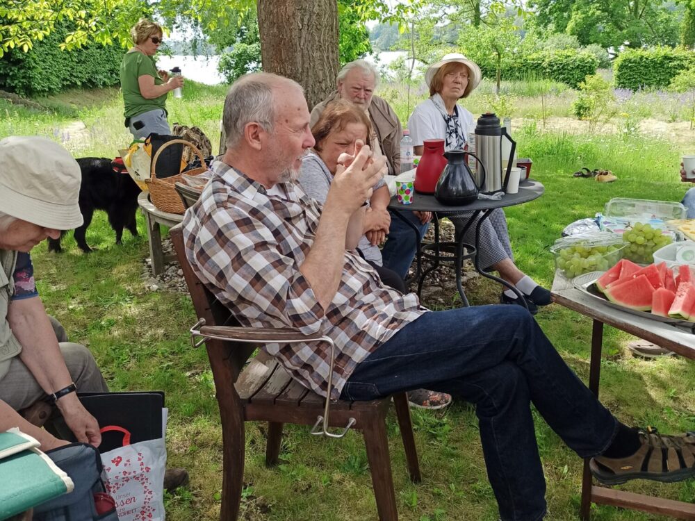 Leute sitzen im Garten unter einem Baum mit Kaffee und Melonenstücken.