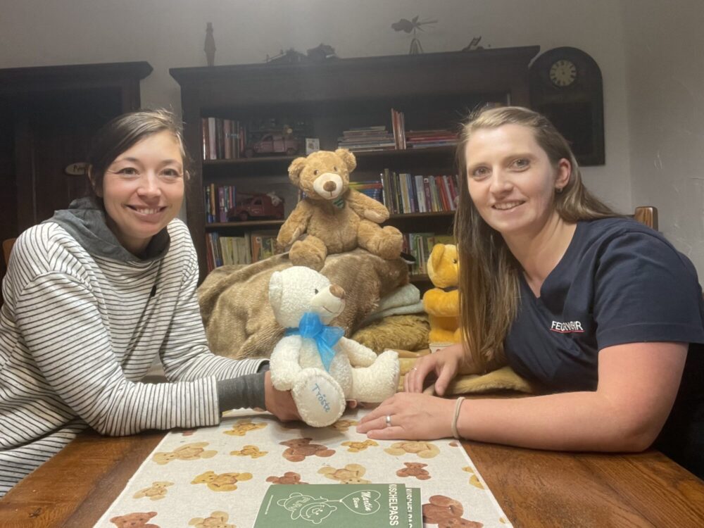 Zwei lächelnde Frauen vor einem Bücherregal mit Teddybären auf dem Tisch.