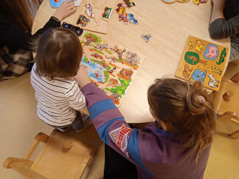 Eine Frau spielt mit einem Kleinkind mit Holzspielzeug am Tisch, von oben fotografiert.