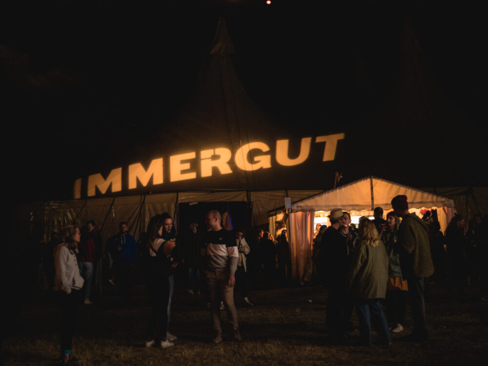 Leute stehen in Grüppchen zusammen in der Dunkelheit vor einem Zelt mit einem beleuchteten Schriftzug darüber.