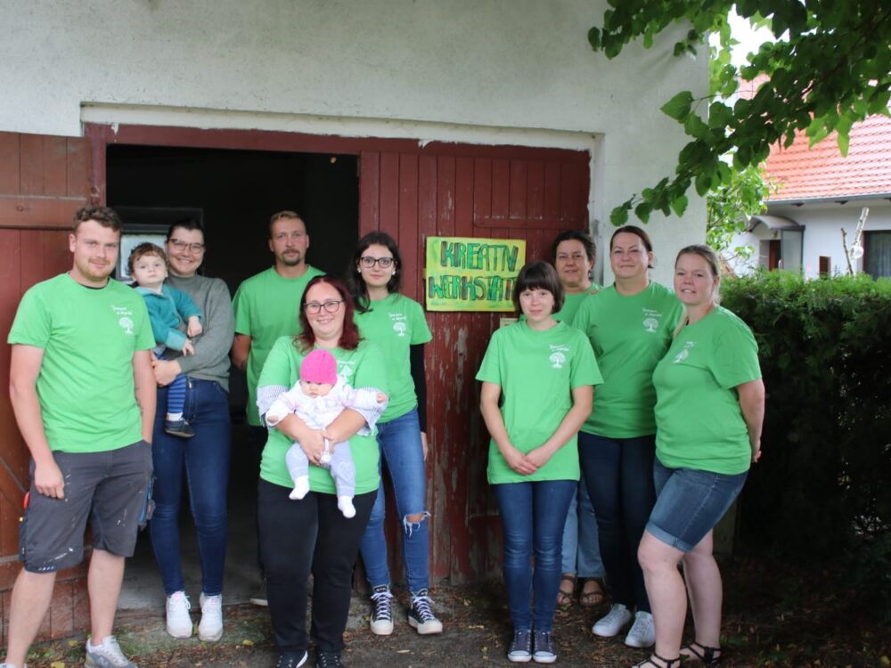 Menschen in grünen T-Shirts stehen lächelnd vor einer roten Garage unter einem Baum. Baum,