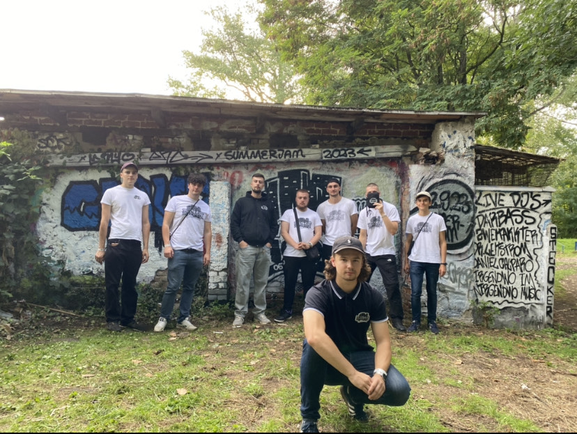Junge Männer in weißen und schwarzen Shirts stehen vor einem grafittiverzierten, älteren Gebäude, im Vordergrund ein junger Mann im schwarzen T-Shirt und Basecap.