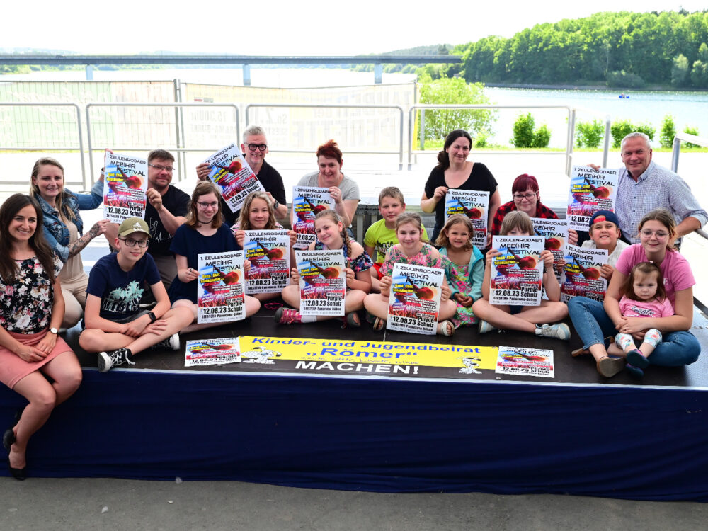 Kinder und Erwachsene sitzen auf einer Bühne am See und halten Plakate hoch “Zeulenrodaer Mee(h)r Festival”.