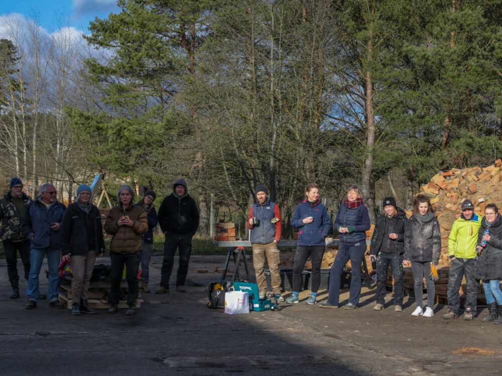 Gruppe in Arbeitssachen steht auf einer Baustelle im Grünen neben einem Haufen Ziegel und macht Pause.