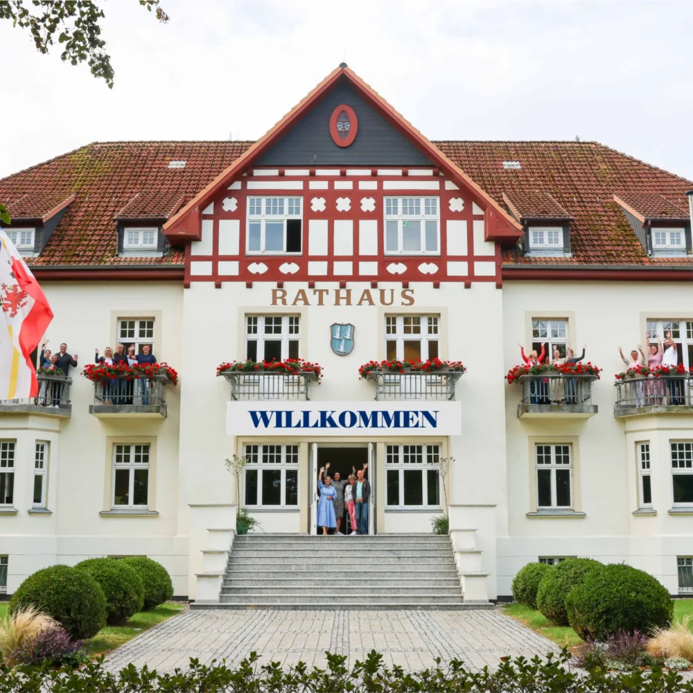 Weißes Gebäude mit Rathaus und Willkommen-Schildern, winkende Menschen am Eingang, Fahnen an den Seiten.