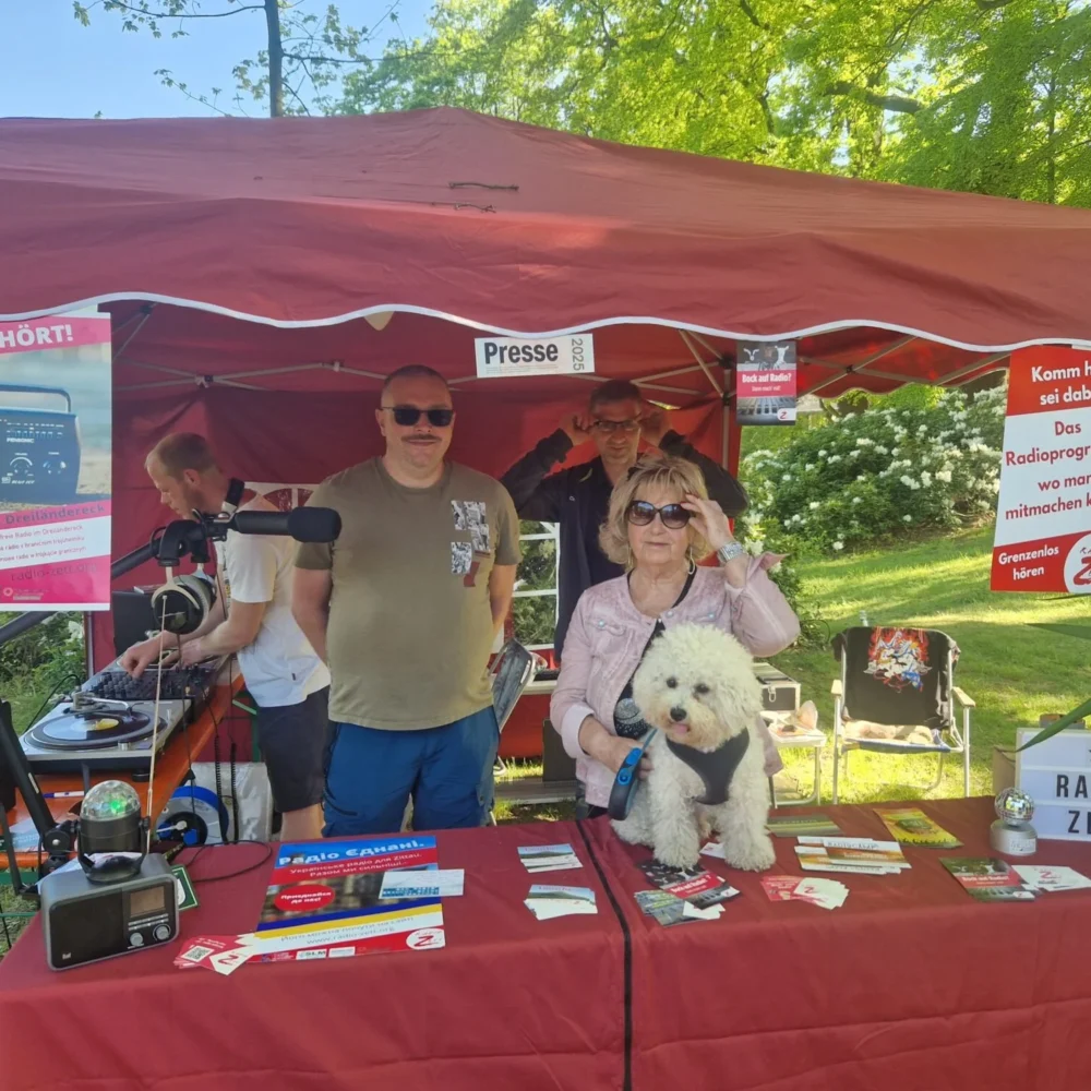 Drei Personen und ein flauschiger weißer Hund an einem Radiostand unter einem roten Zelt.