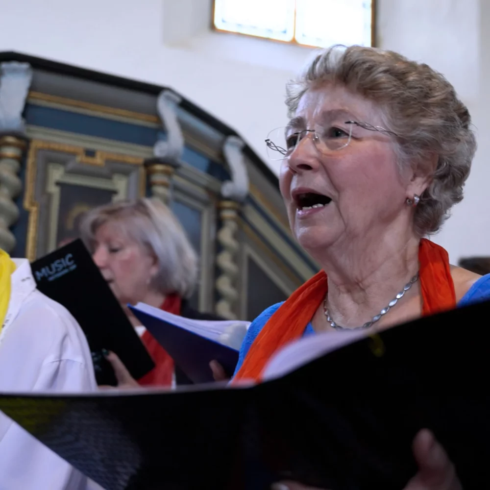 3140_Foto6_mmpro media AG_David Katz Ältere Frau singt in einem Chor in einer Kirche, hält Notenblätter, weitere Chormitglieder im Hintergrund, Tageslicht durch Fenster.